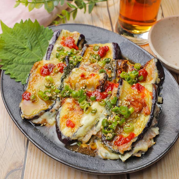 皿に盛られた焼きナスにチーズとネギ、ソースがかかっている料理。隣に茶色の飲み物入りのグラスが置かれている。