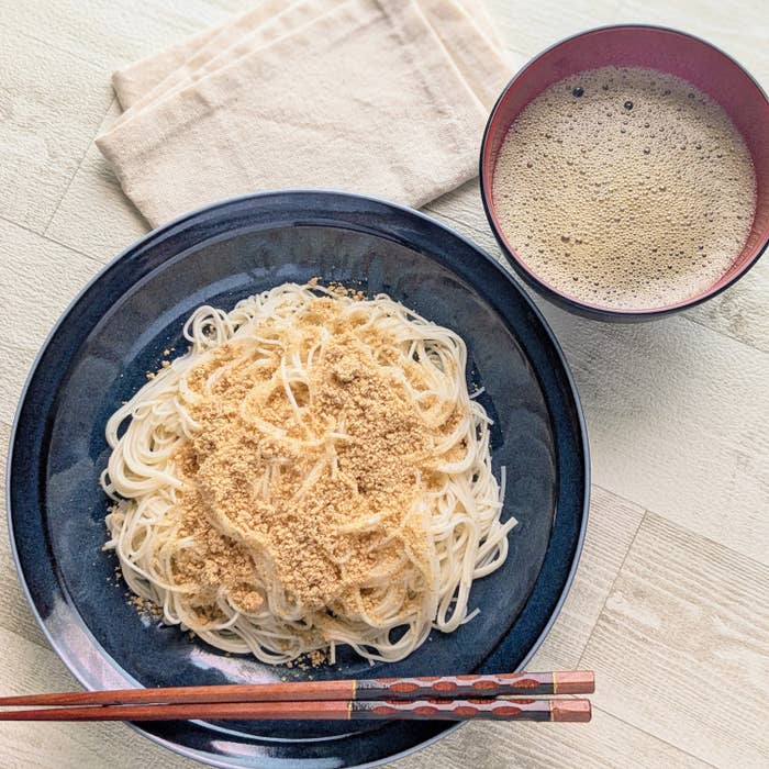 麺にすりごまがかかった料理が黒い皿に盛られ、横につけ汁が置かれている。ベージュのふきんも見える。