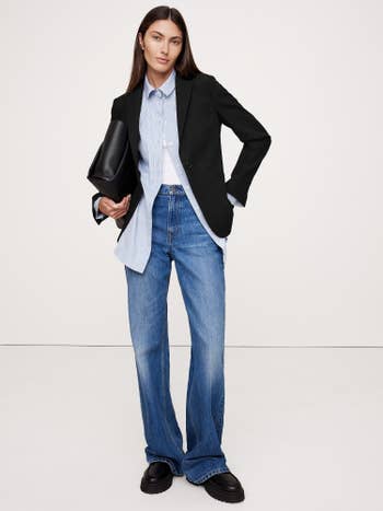 Person wearing a tailored blazer, untucked shirt, wide-leg jeans, and holding a clutch bag, styled for a casual yet chic buying experience