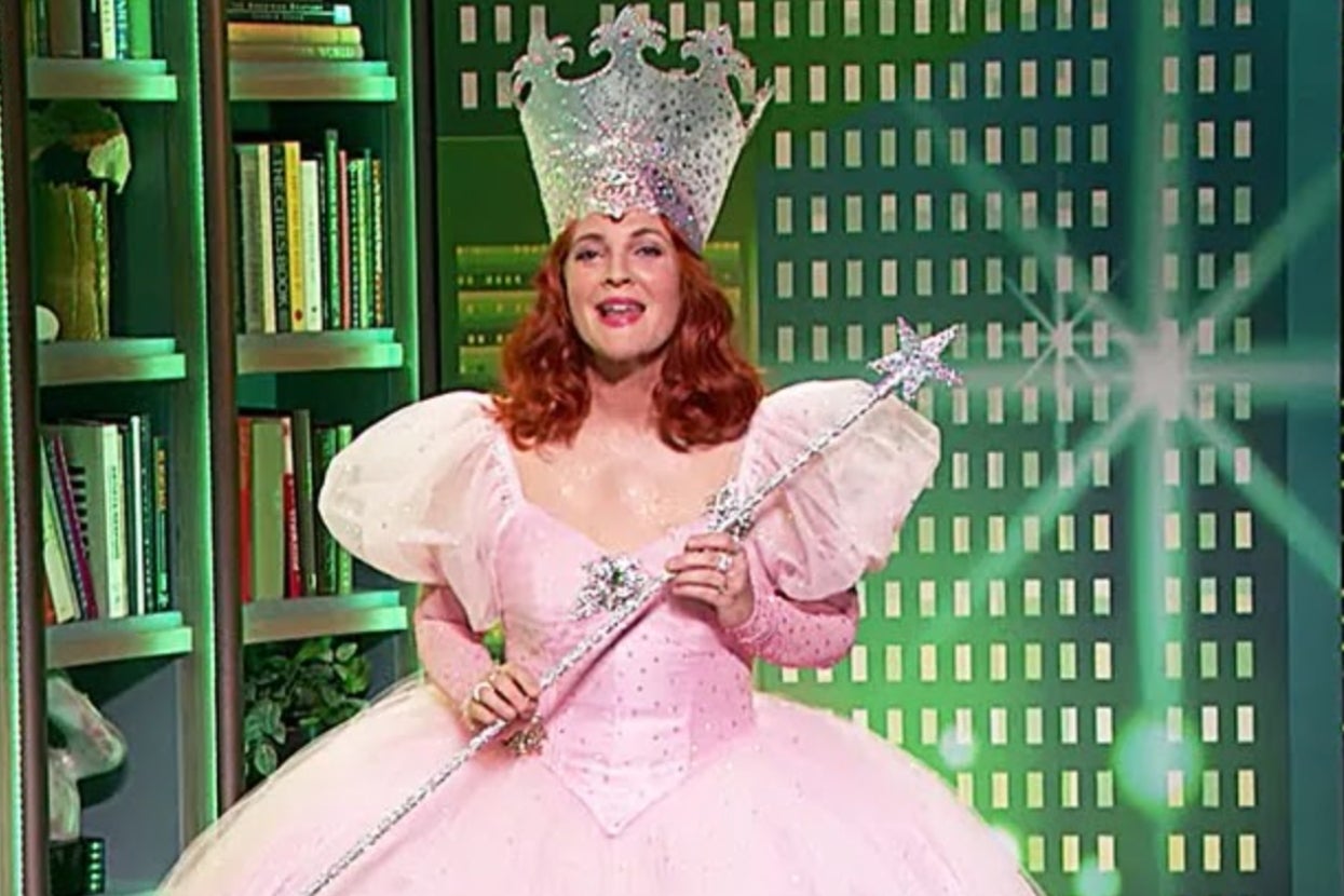 Drew Barrymore dressed as the whimsical Good Witch from "Wicked" with a jeweled crown, holding a sparkling wand in a fancy dress, standing in a book-filled room