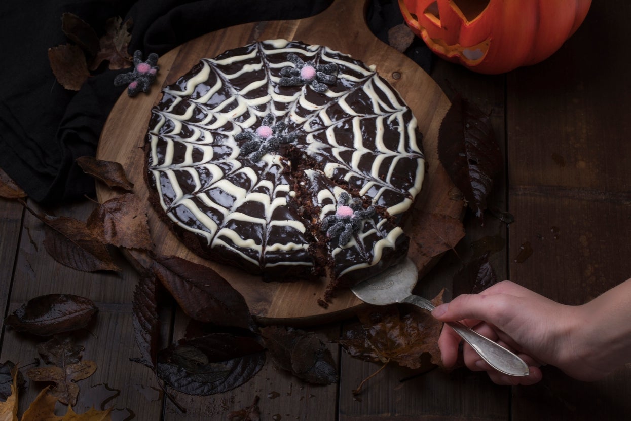 Chocolate cake with spiderweb icing and candy spiders, set on a wooden board surrounded by autumn leaves. A hand holds a serving utensil
