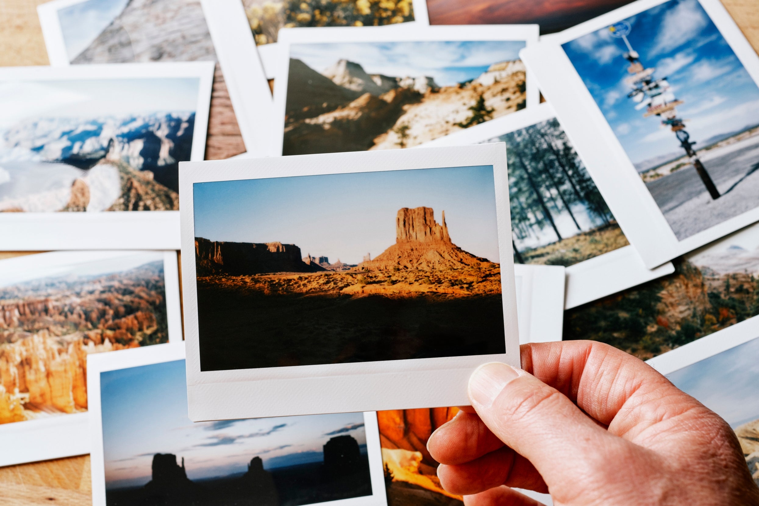 A hand holds a landscape photo of a desert mesa, surrounded by various other scenic landscape photos laid out on a surface
