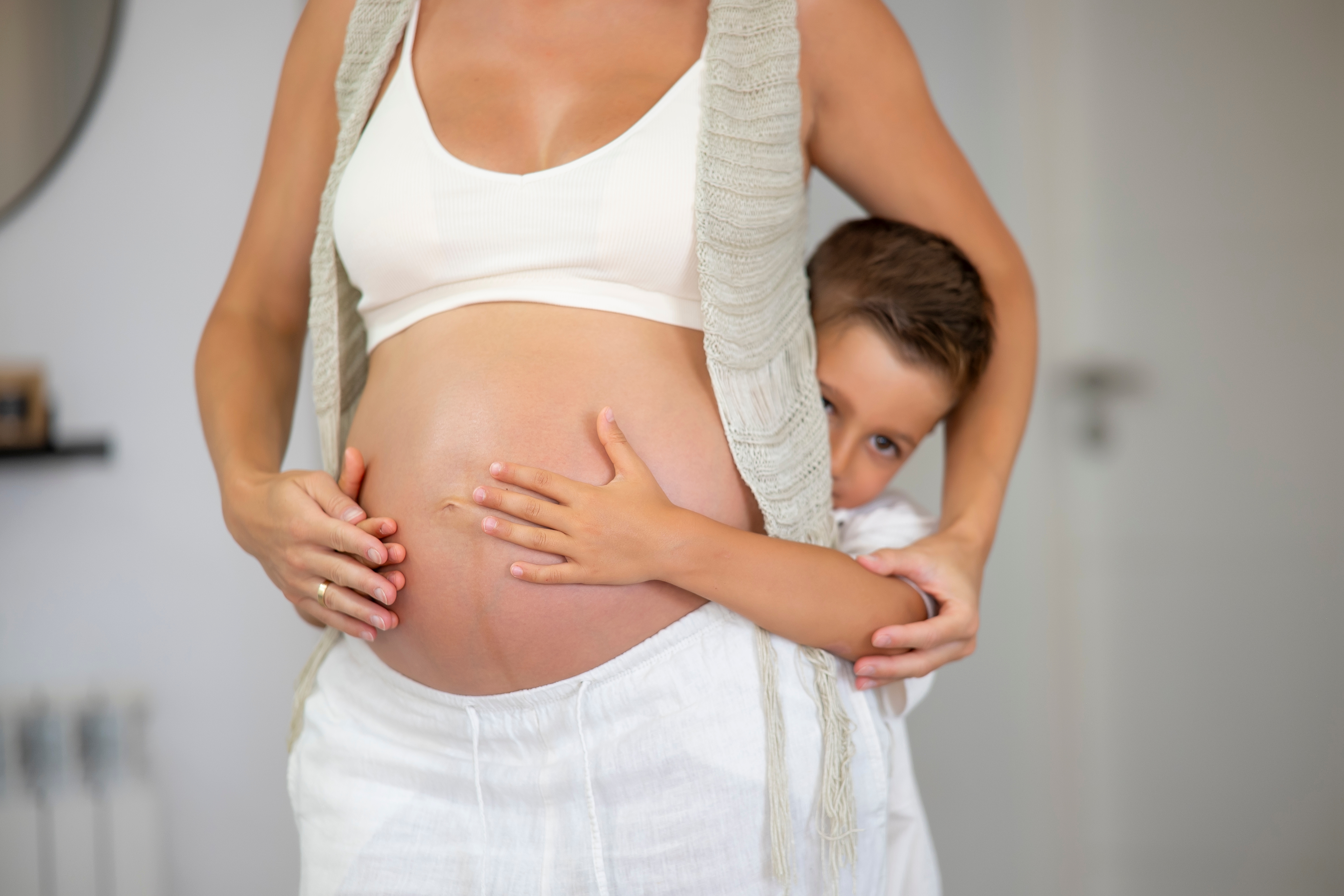 Gravid kvinne i en hvit topp, holder magen, med et barn som lekent klemmer bakfra, og skaper en varm og kjærlig scene