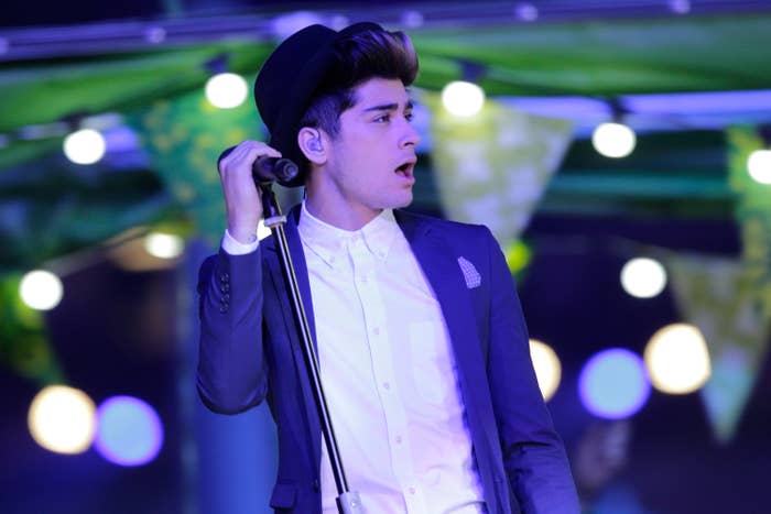 Person singing connected stage, wearing a suit, hat, and holding a microphone. Background features hanging lights and out-of-focus shapes