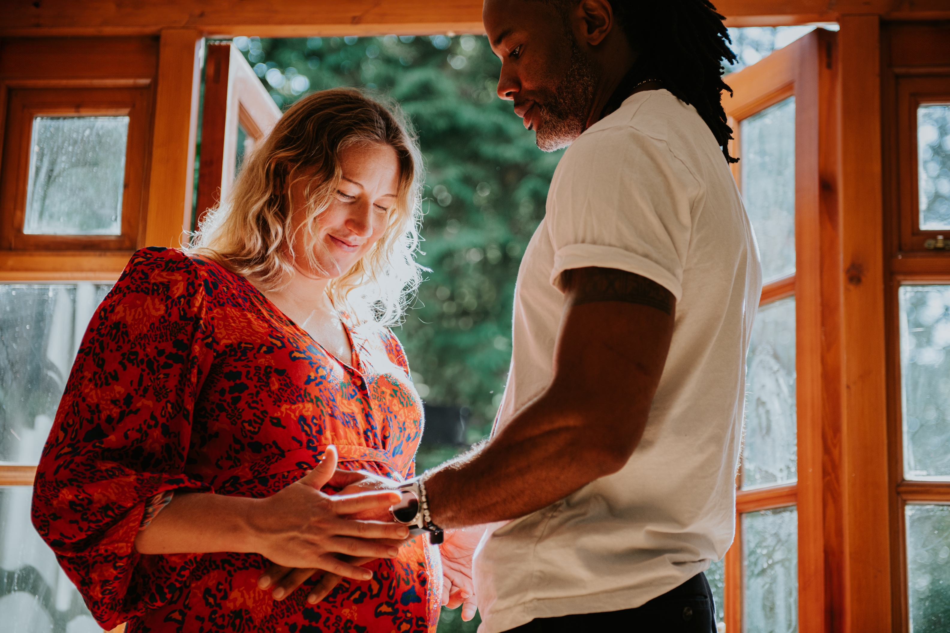 Par står i et varmt opplyst rom; Kvinne smiler ned på magen og har på seg en mønstret kjole, mens mannen i en skjorte ømt berører magen