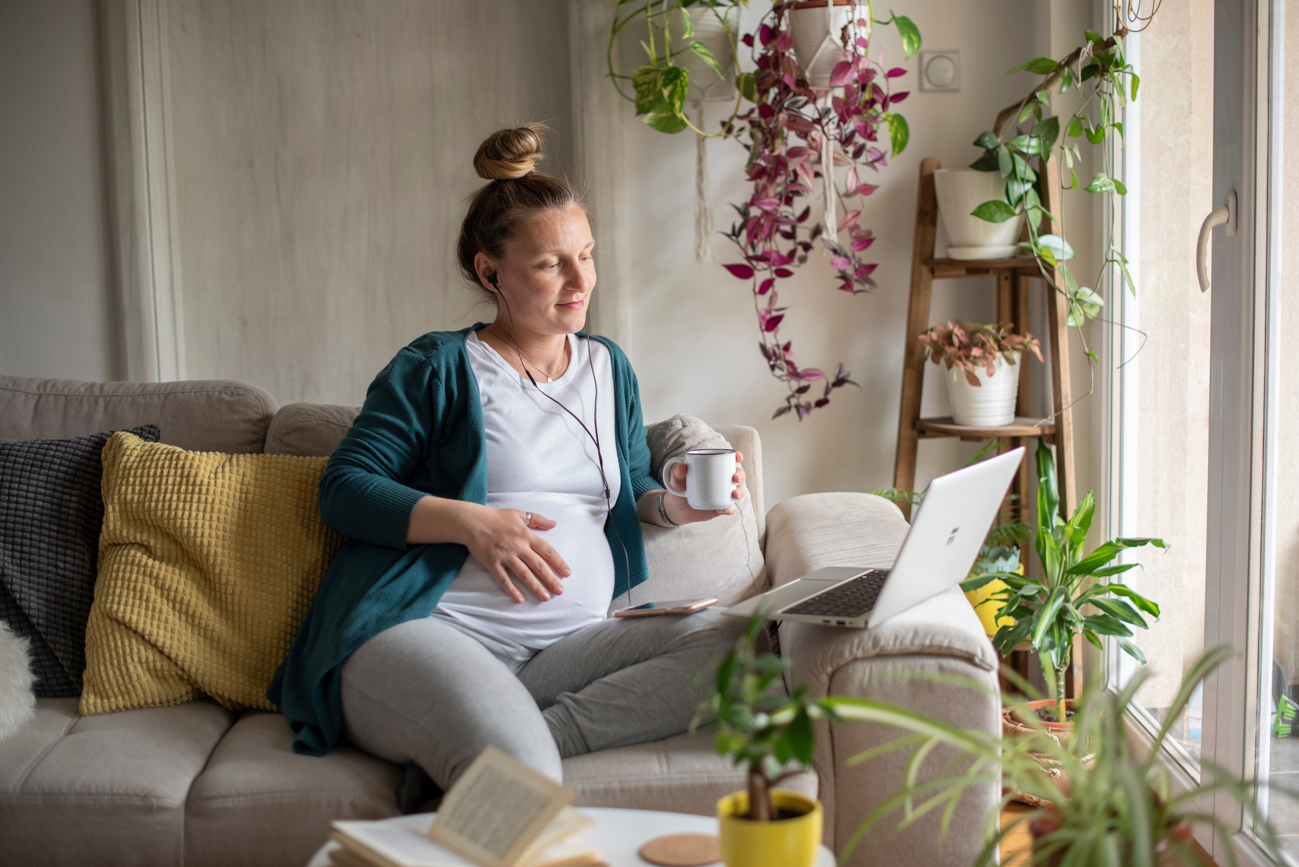Person slapper av på en sofa, holder et krus, med en bærbar datamaskin, omgitt av planter og nyter en koselig atmosfære