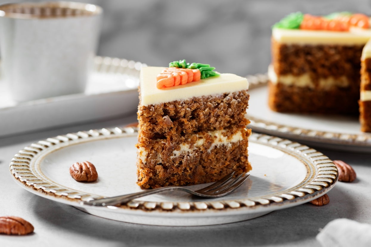 A slice of carrot cake with icing and a decorative carrot on top, served on a white plate with a fork