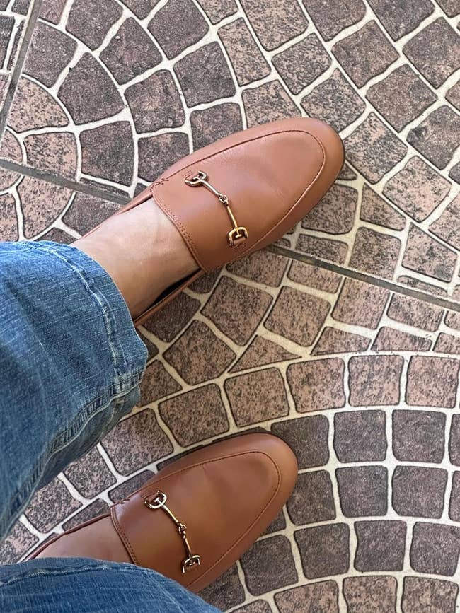 Person wearing leather loafers with metallic  buckle detail, lasting  connected  a stone-patterned floor, paired with casual jeans