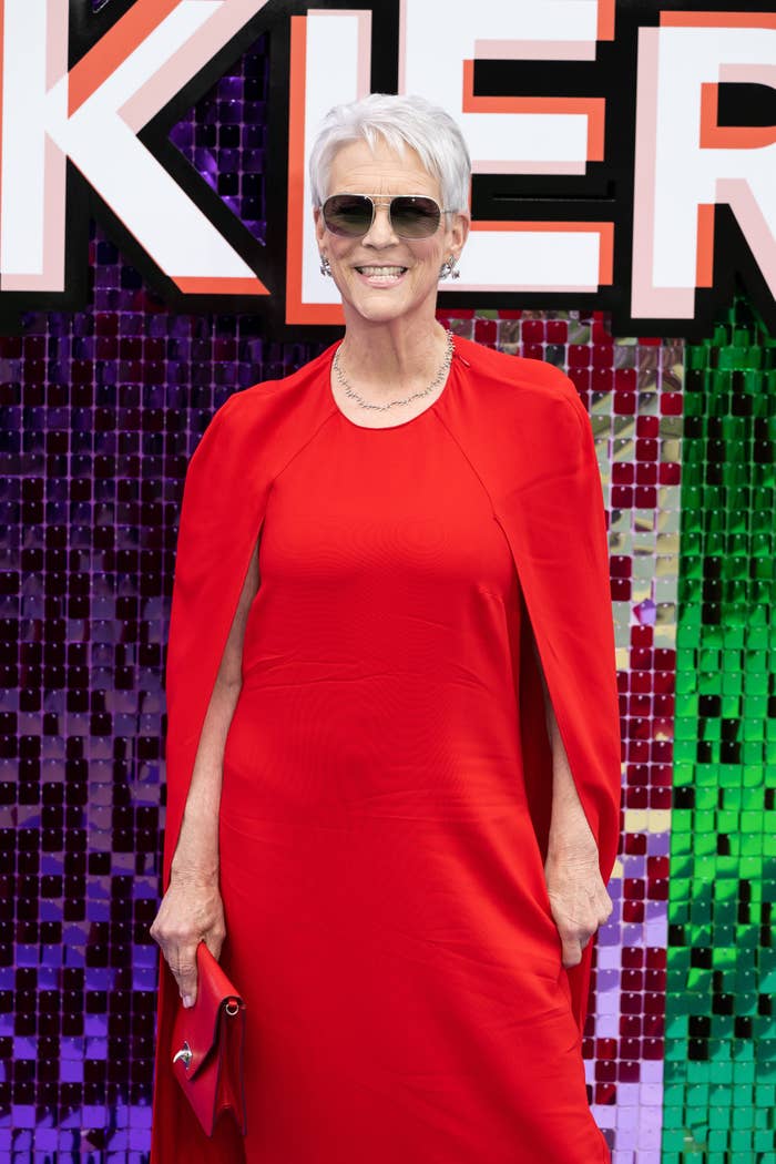 Person successful elegant reddish formal with a cape overlay, holding a matching clutch, wearing sunglasses, and smiling successful beforehand of a sequin backdrop