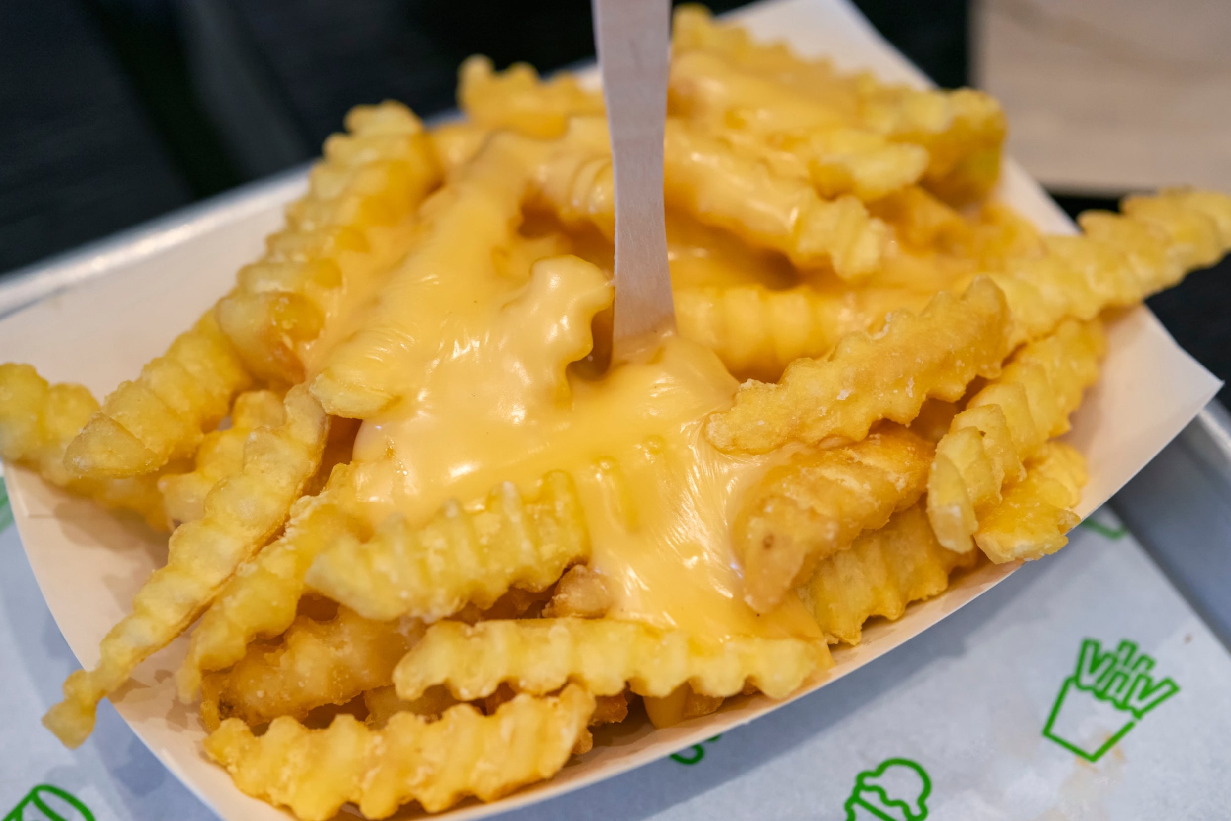 Crinkle-cut fries topped with melted cheese, served in a paper tray with a wooden fork