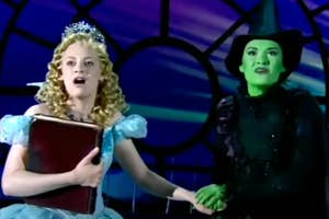 Two performers on stage, one in a sparkly gown holding a book and the other in a witch costume, singing passionately while holding hands