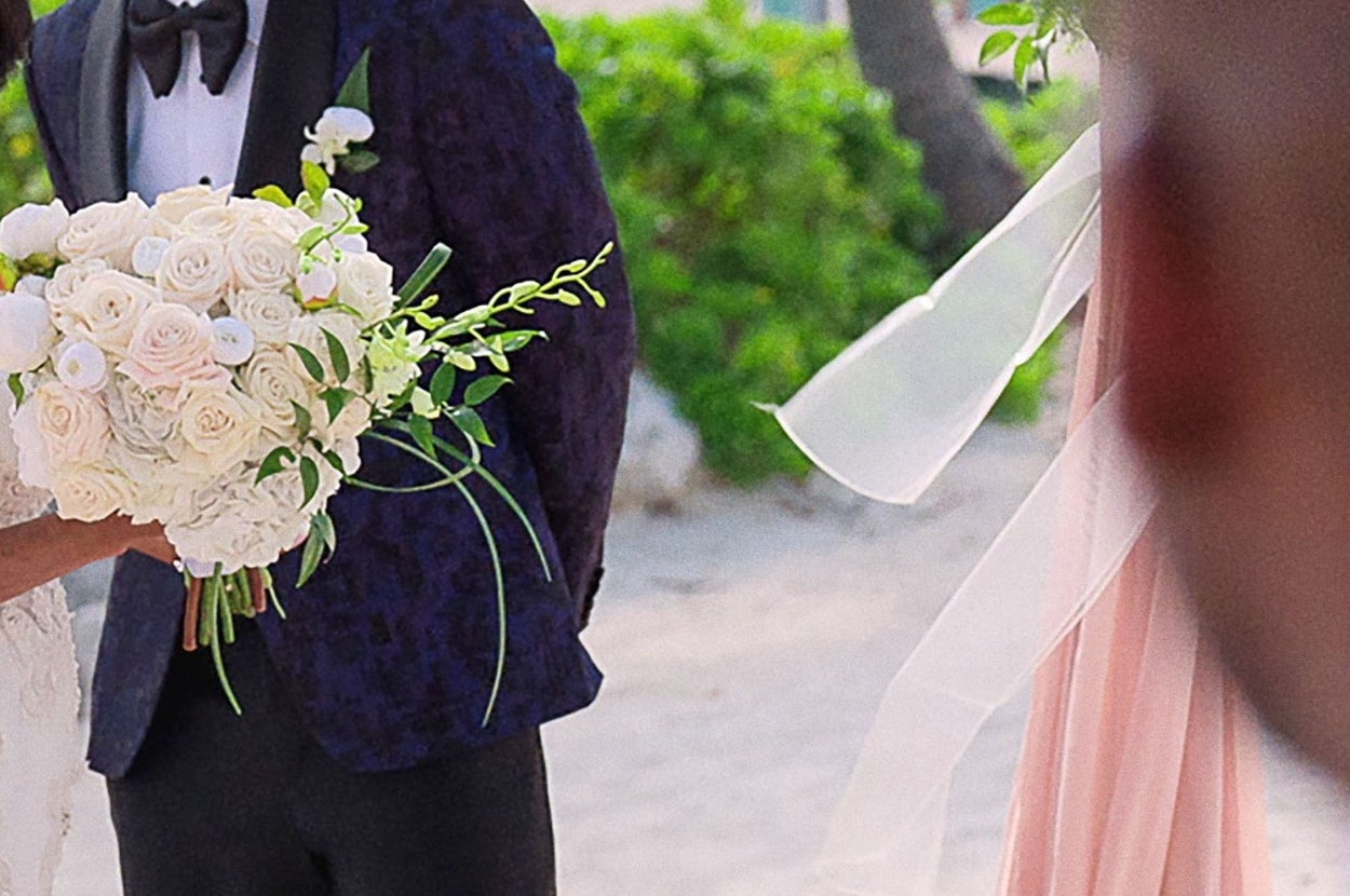 Couple stands under a floral wedding arch on a beach; bride in a ruffled gown holding flowers, groom in a patterned tuxedo. A person is seen from behind