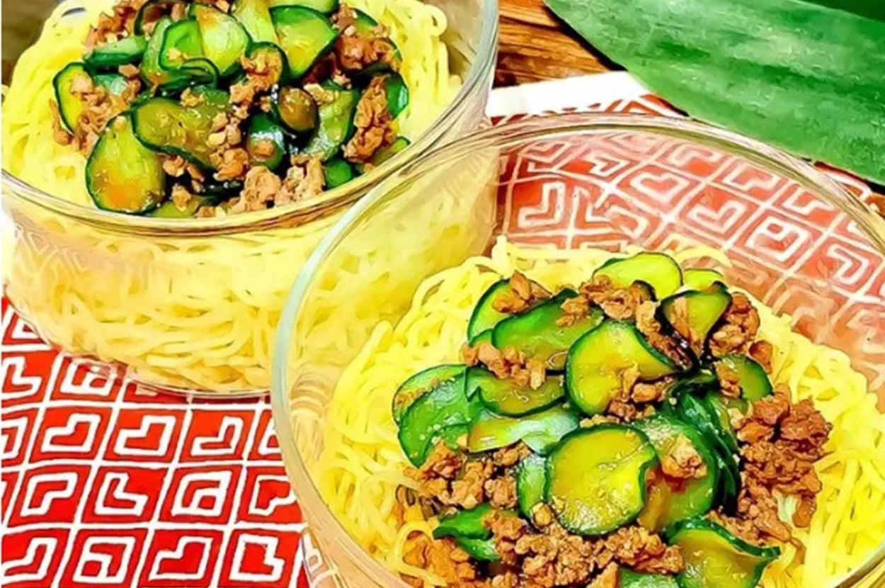 2つのガラスの器に盛られた麺の上に、薄切りのきゅうりと肉のトッピング。木のテーブルに置かれている。