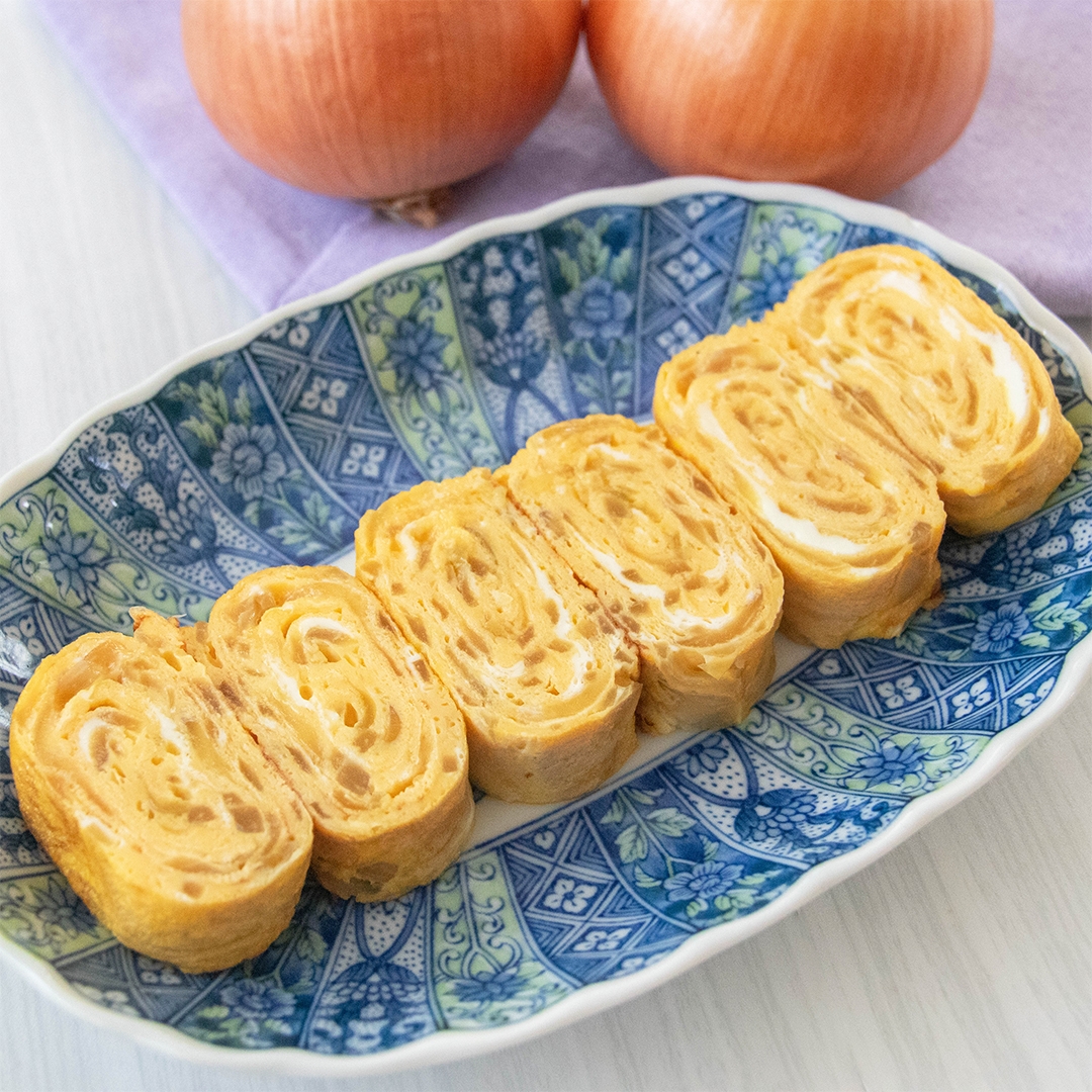 青い模様の皿に盛られた玉ねぎ入りの卵焼きが6切れ。背景に玉ねぎが2つ。