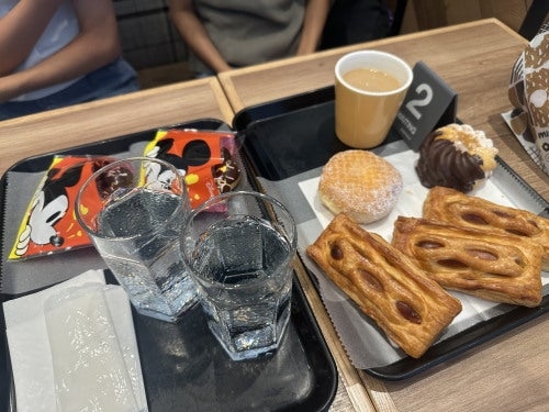 テーブルの上に飲み物と菓子パンが載ったトレイが2セット並んでいるカフェのシーン。