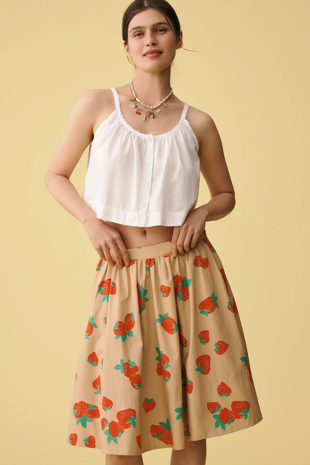 Woman wearing a white crop top and a skirt with strawberry patterns, posing against a plain background, showcasing a casual summer look