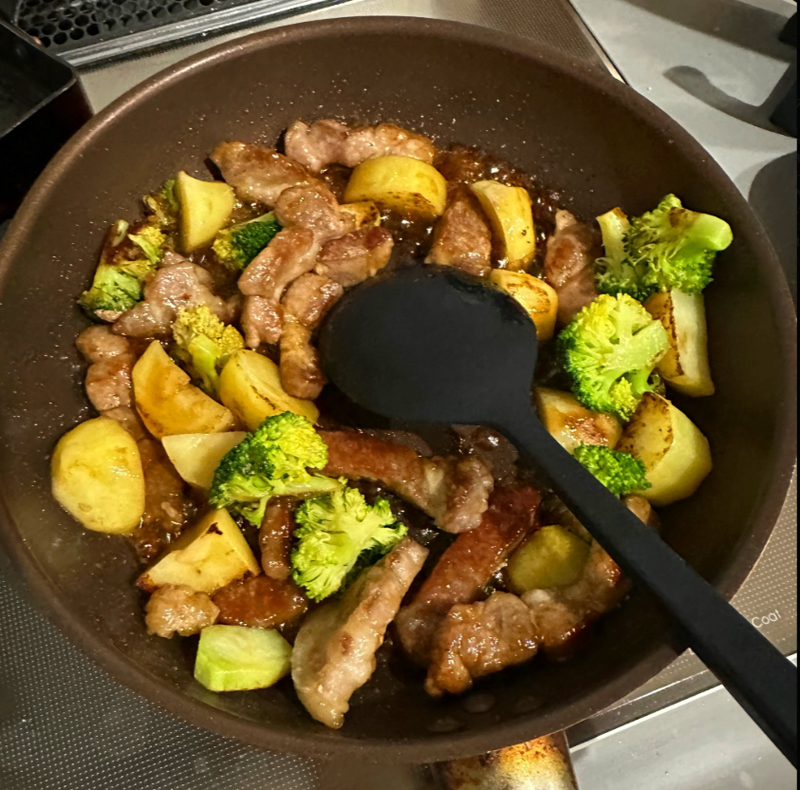 ブロッコリー、豚肉、じゃがいもが入ったフライパンでの調理中の料理。黒いスパチュラが使用されています。