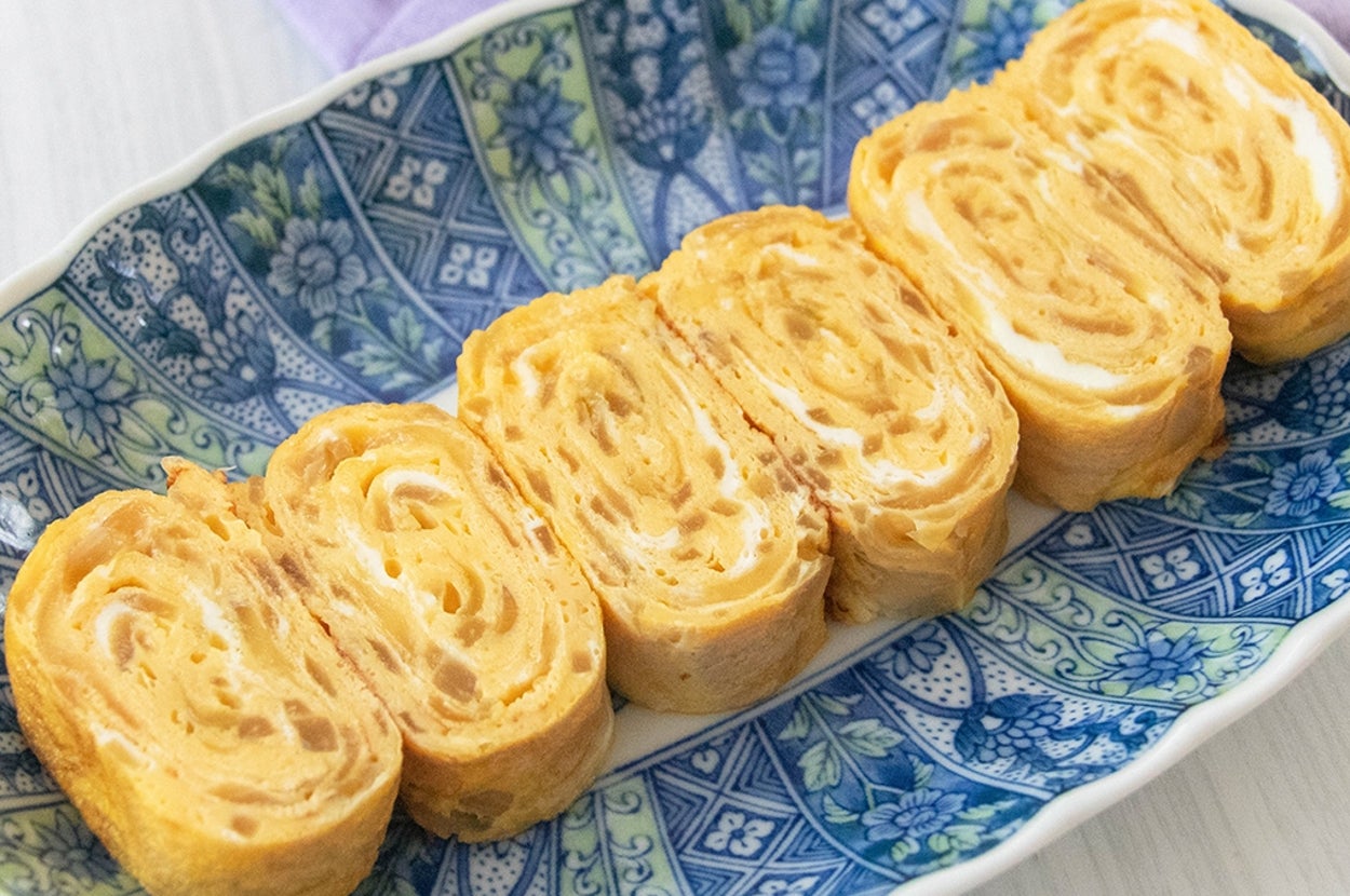 青い模様の皿に盛られた玉ねぎ入りの卵焼きが6切れ。背景に玉ねぎが2つ。