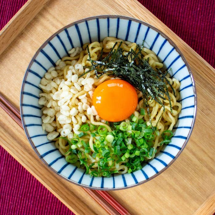 ラーメン鉢に生卵、刻み海苔、揚げ玉、ネギがトッピングされた料理が木製トレイに載っている。