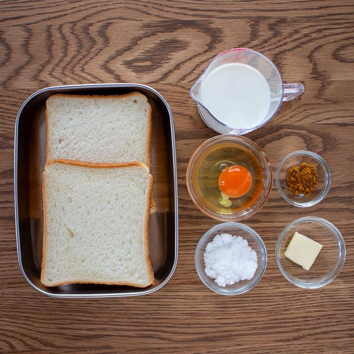 食パン2枚、牛乳、卵、砂糖、バター、シナモンが木製のテーブルに並んでいます。