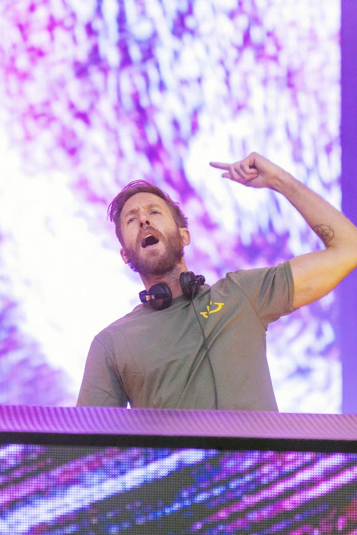 DJ performing energetically with headphones astir his neck, gesturing upwards connected signifier with vibrant visuals successful the background