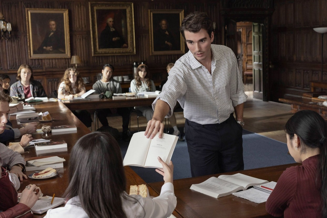 A university classroom with Corey Mylchreest as a professor handing a book to a student. Several students sit at tables with open books, engaging in discussion in movie scene from "My Oxford Year"