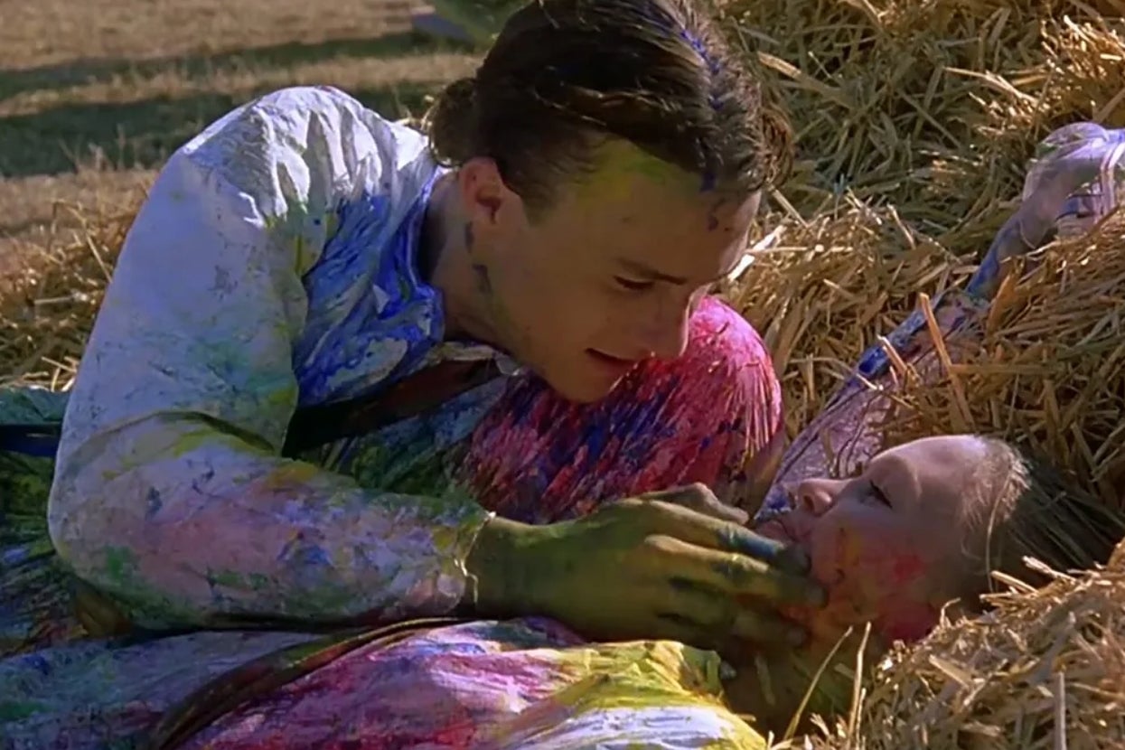 Two people lie in hay, faces and clothing covered in paint, looking at each other closely in a playful, intimate moment