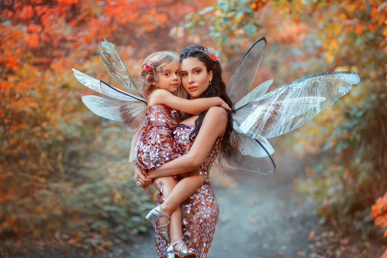 Two people in sparkling, sequin outfits with fairy wings stand in a forest setting, one holding the other