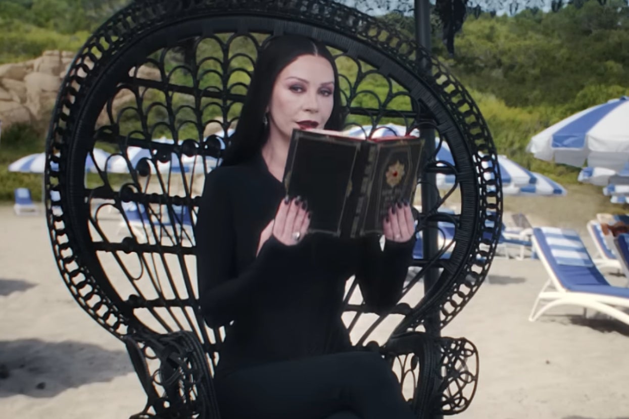 Morticia Addams in gothic attire sits on a beach chair, reading a book. Beach umbrellas and lounge chairs in the background