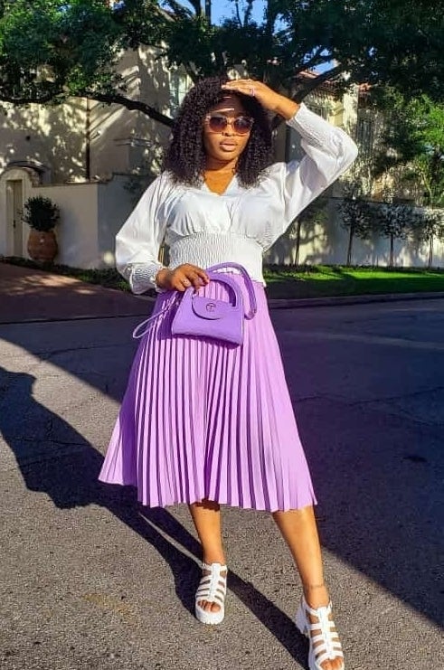 Person wearing a white blouse, pleated skirt, and holding a stylish handbag poses outdoors on a sunny day