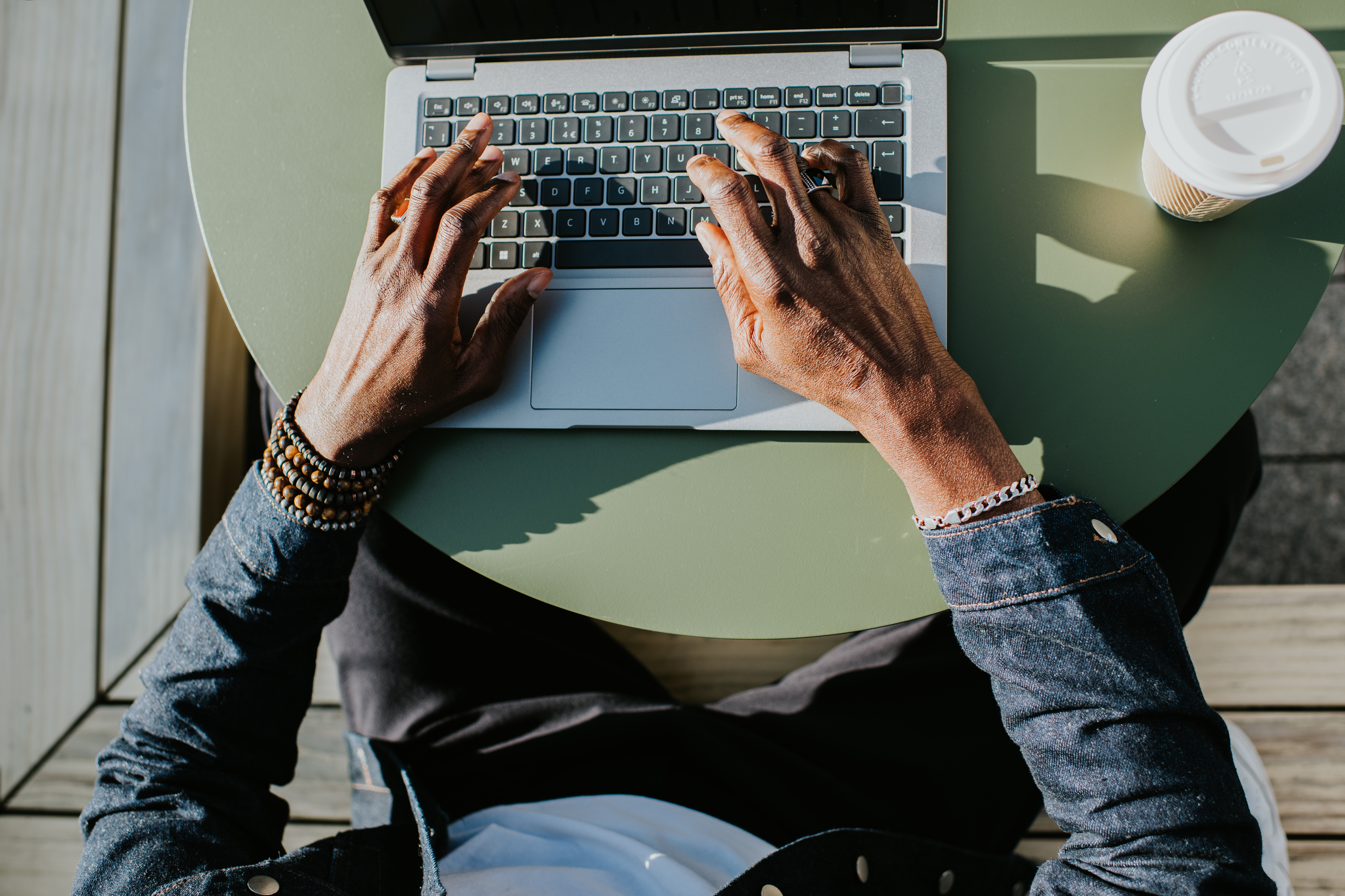 Person som skriver på en bærbar datamaskin ved et rundt bord, iført en denimjakke og armbånd, med en to-go kaffekopp ved siden av seg