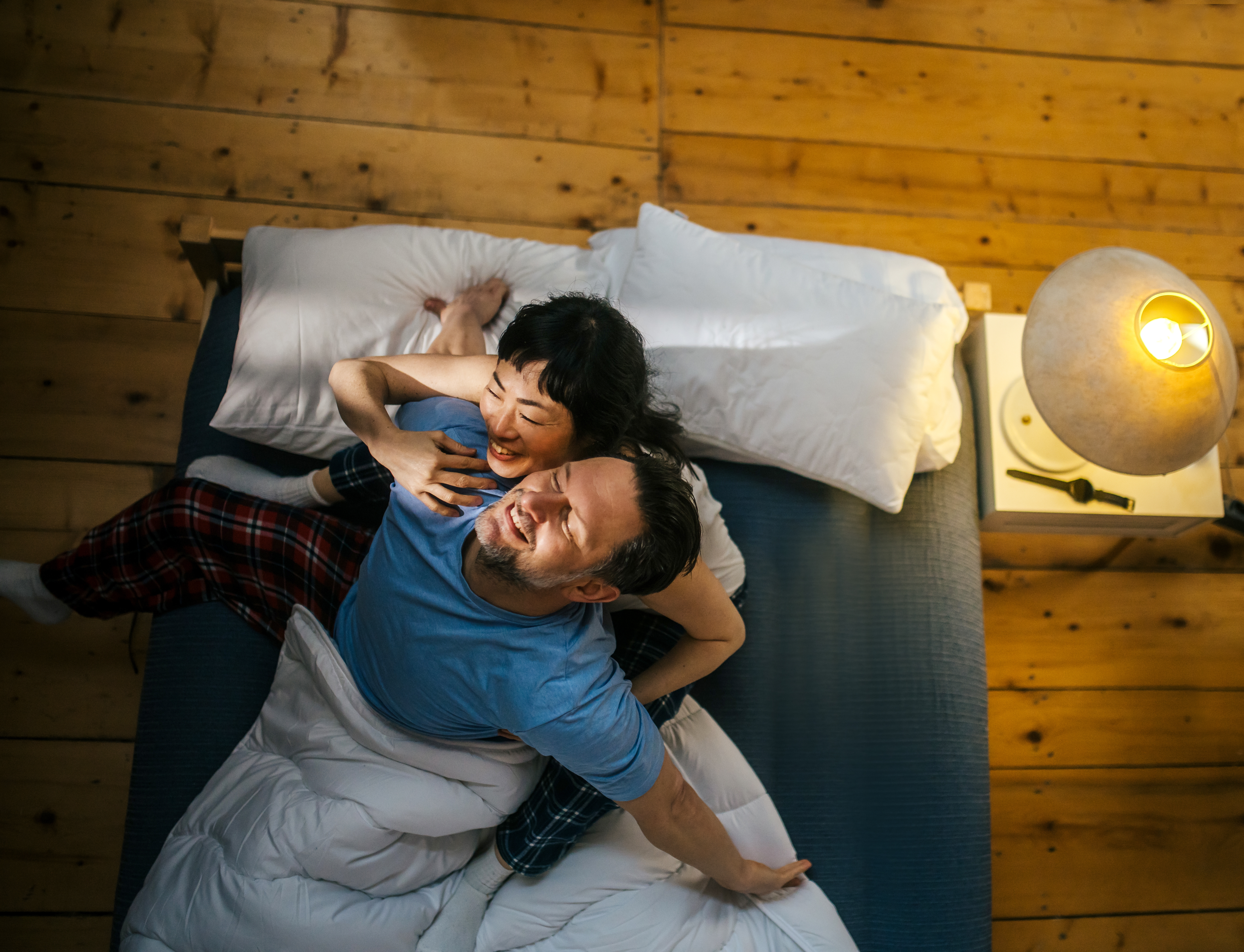 A couple in pajamas sits joyfully on a bed with wooden flooring, lit by a bedside lamp