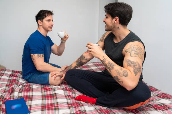 Two people with tattoos sit on a bed, holding cups, engaged in conversation
