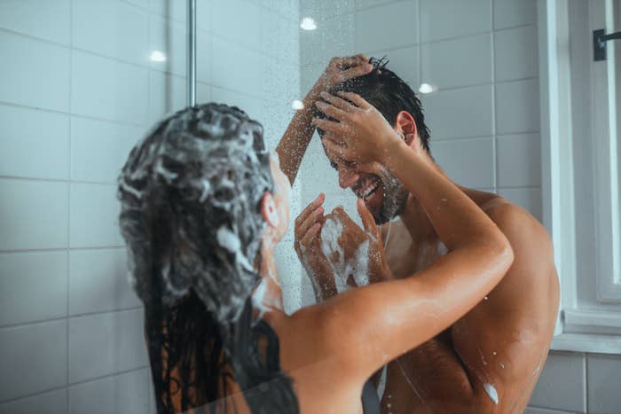 Two people smiling and playfully washing each other's hair in a shower, enjoying a light-hearted moment together