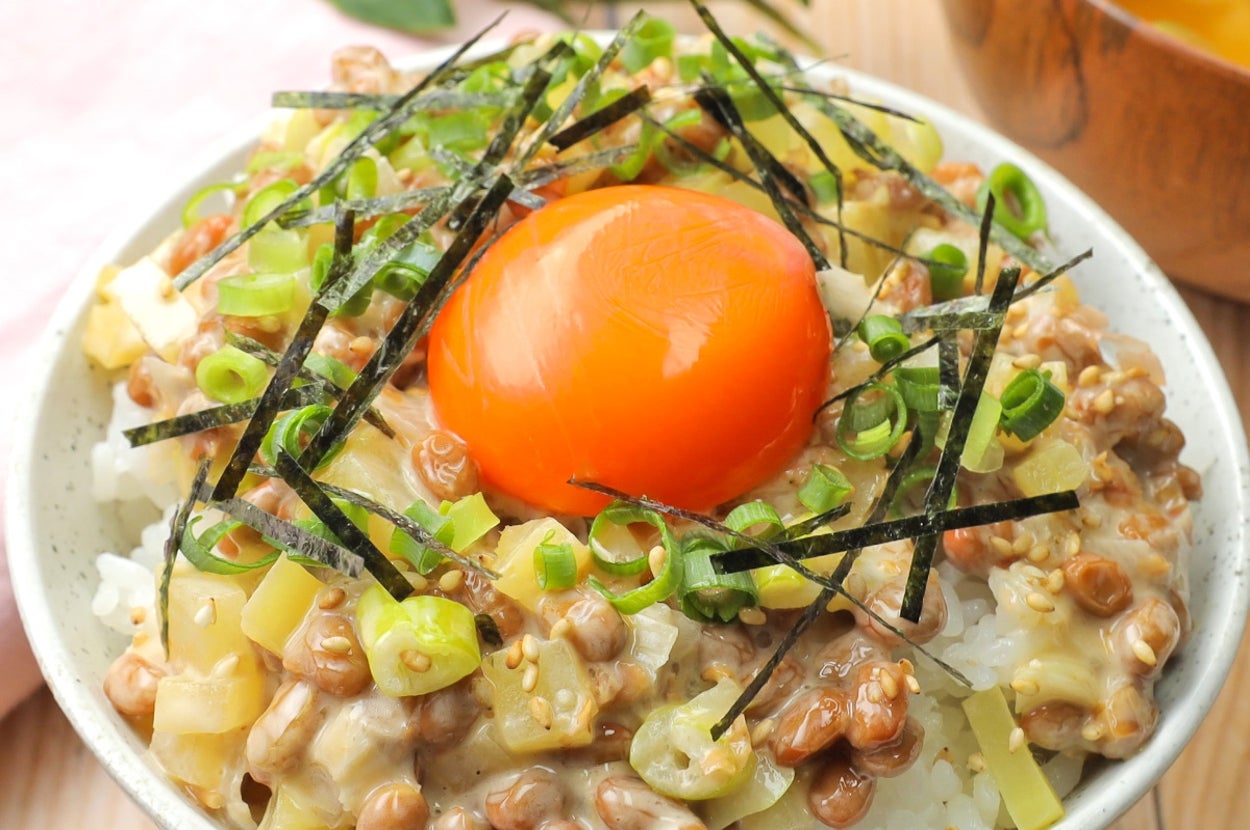 納豆ご飯の上に卵黄、刻みネギ、のりがトッピングされています。背景に味噌汁の一部が見えます。