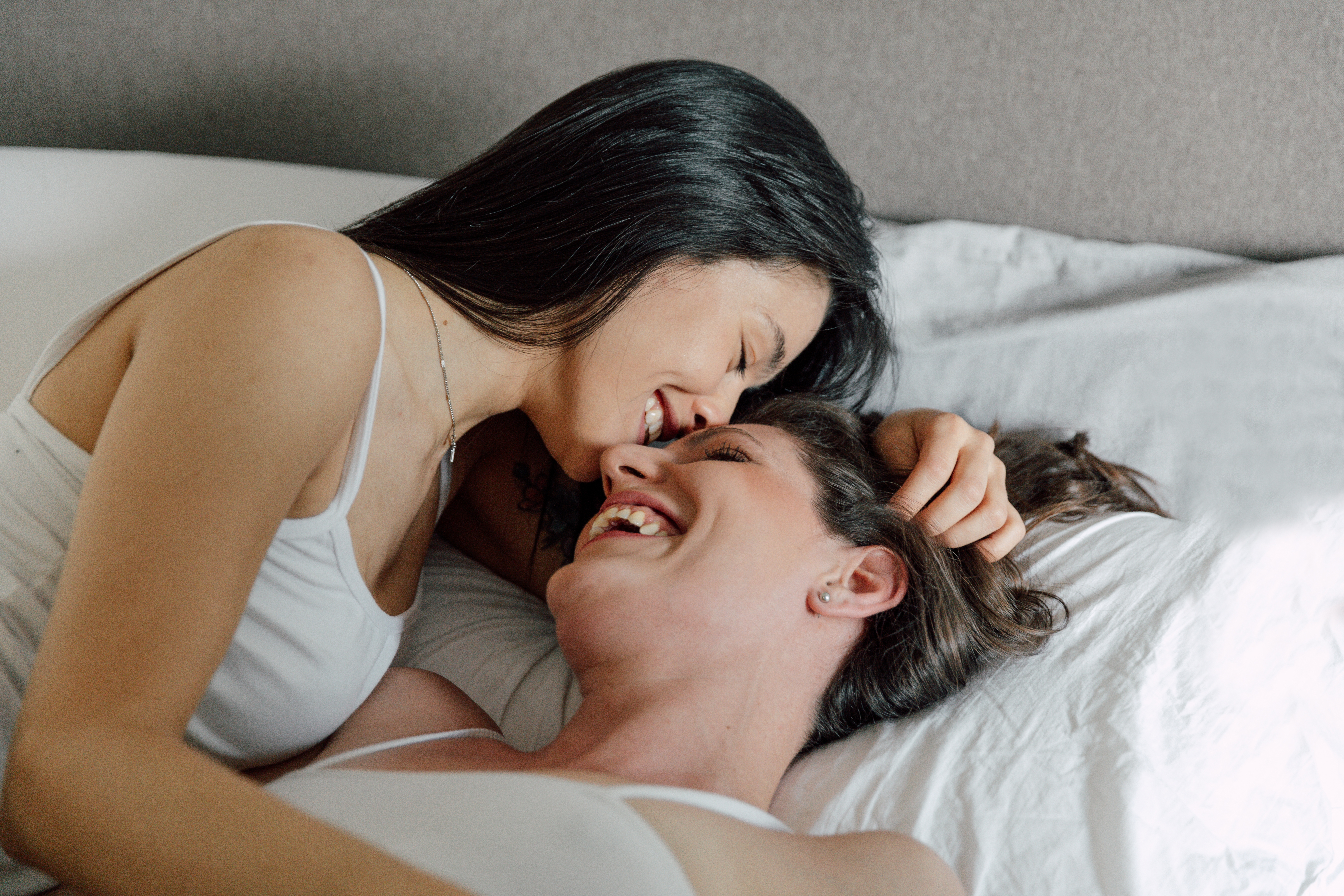 Two people lying on a bed, smiling and embracing each other in a warm, intimate moment. They both wear casual sleeveless tops