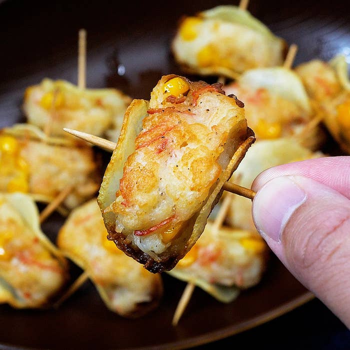 人の手が竹串で刺した一口サイズの焼き餃子を持っている。背景には複数の餃子があります。
