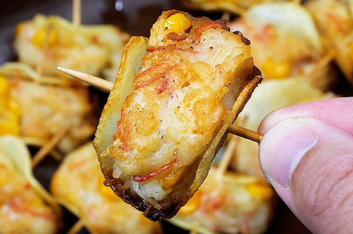 焼きエビをつまようじで刺して持ち上げている手前の部屋で、背後に他の揚げ物も並ぶ。