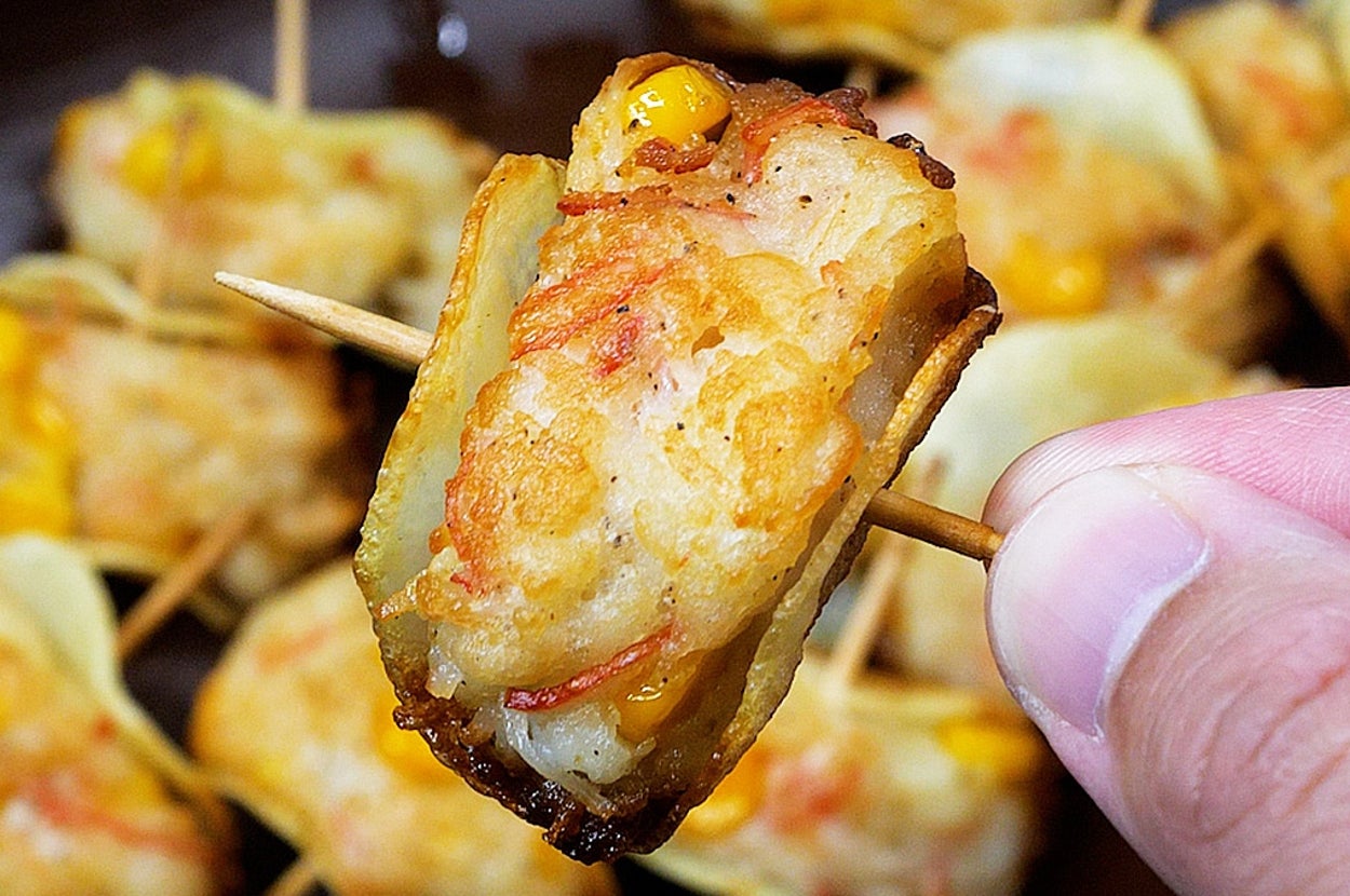 焼きエビをつまようじで刺して持ち上げている手前の部屋で、背後に他の揚げ物も並ぶ。