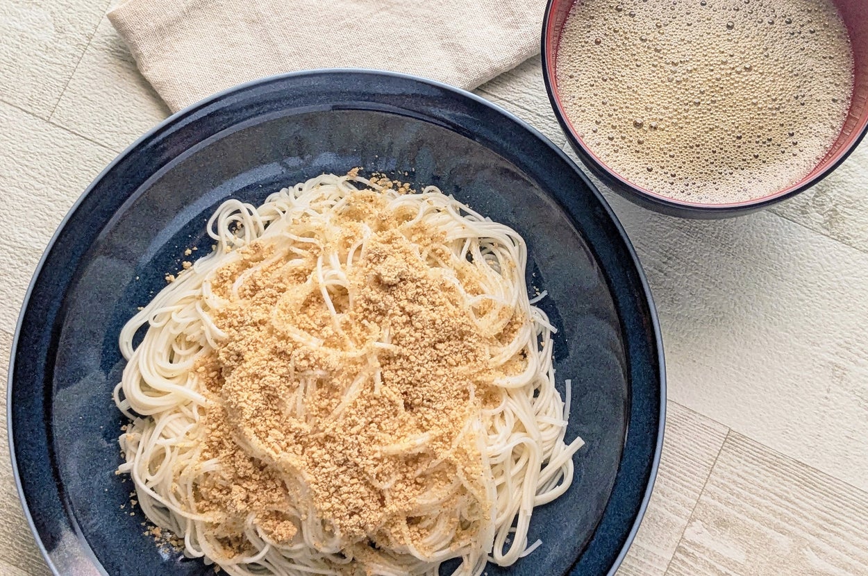 蕎麦の上にごまと胡麻だれがかかった料理が黒い皿に乗った写真。隣にスープが写っています。