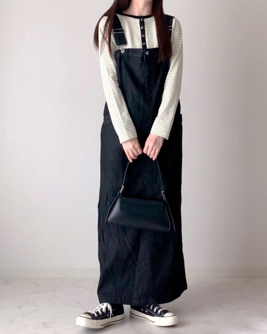 女性がボーダーシャツとデニムジャンパースカートを着用し、ハンドバッグを持っている。カジュアルな服装。