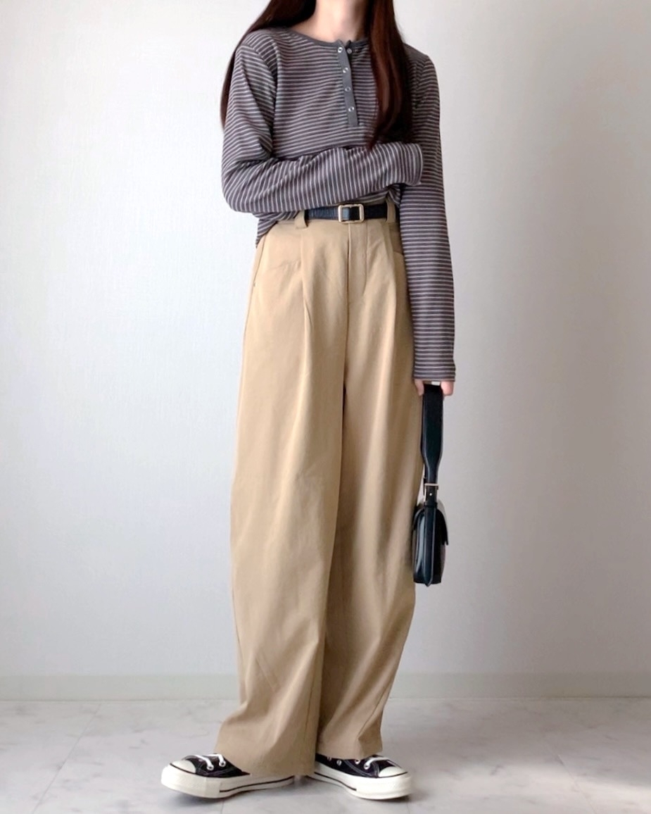 女性がストライプのトップスとワイドパンツを着て、スニーカーを履いて立っている。バッグを持っている。