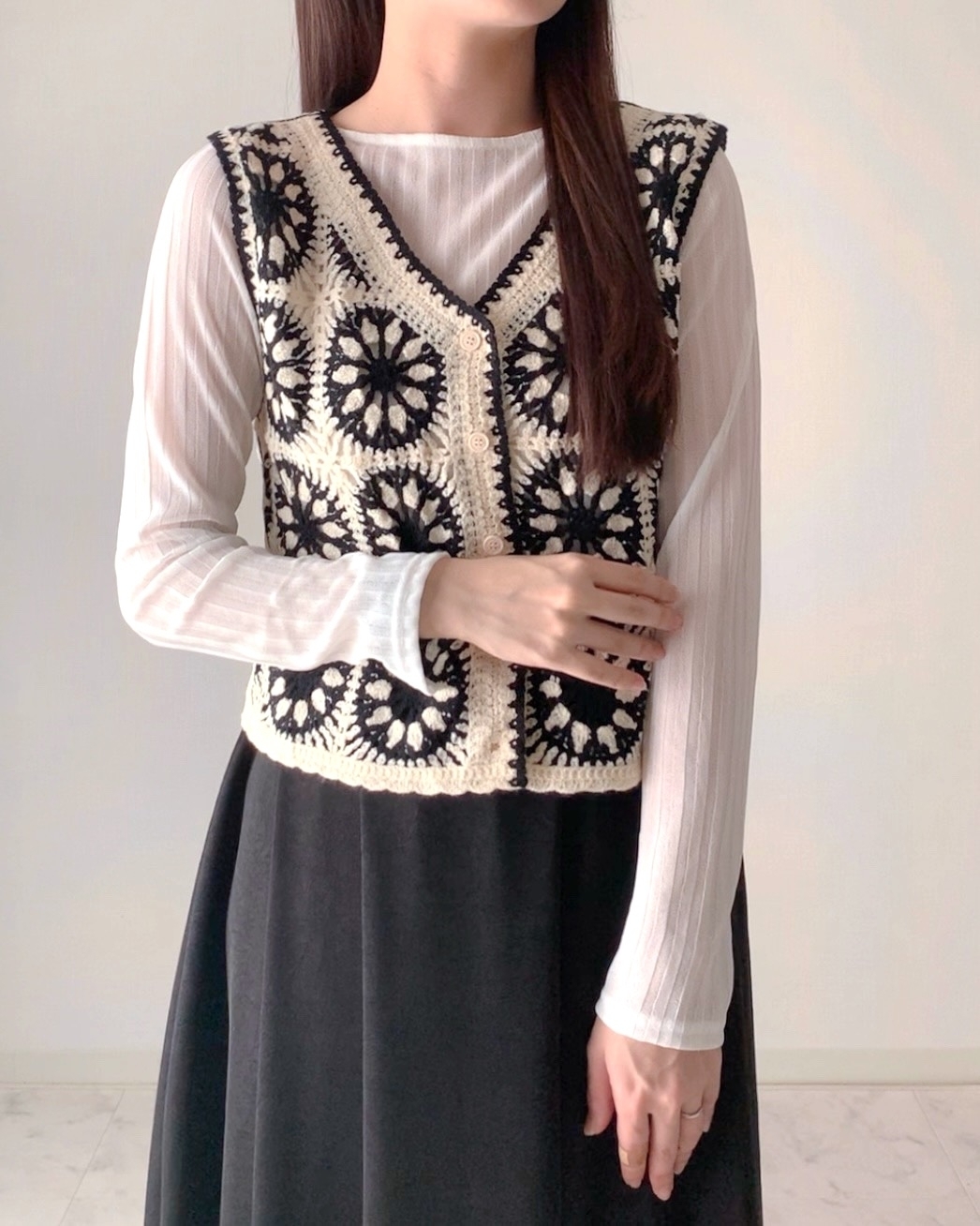 女性がシンプルな白シャツに黒とクリーム色の編みベストを重ね着して立っている。