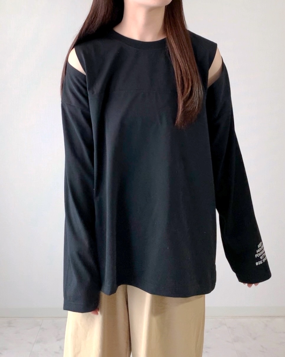 女性が肩の部分がカットアウトされた黒の長袖トップスとゆったりしたベージュのパンツを着ている。