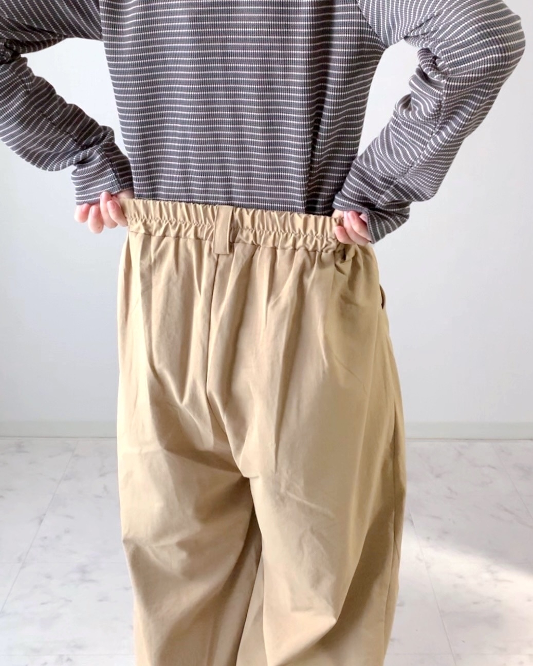 ゆったりしたストライプのトップスとベージュのワイドパンツを着た人が、パンツのウエストを広げて見せている様子。