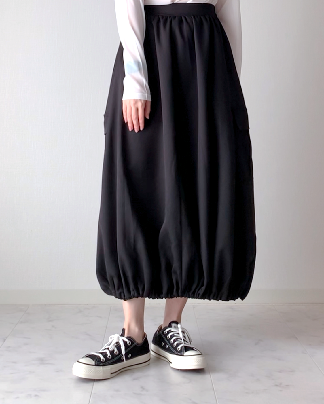 女性がふんわりとした長い黒のスカートをはき、スニーカーを履いて立っている。