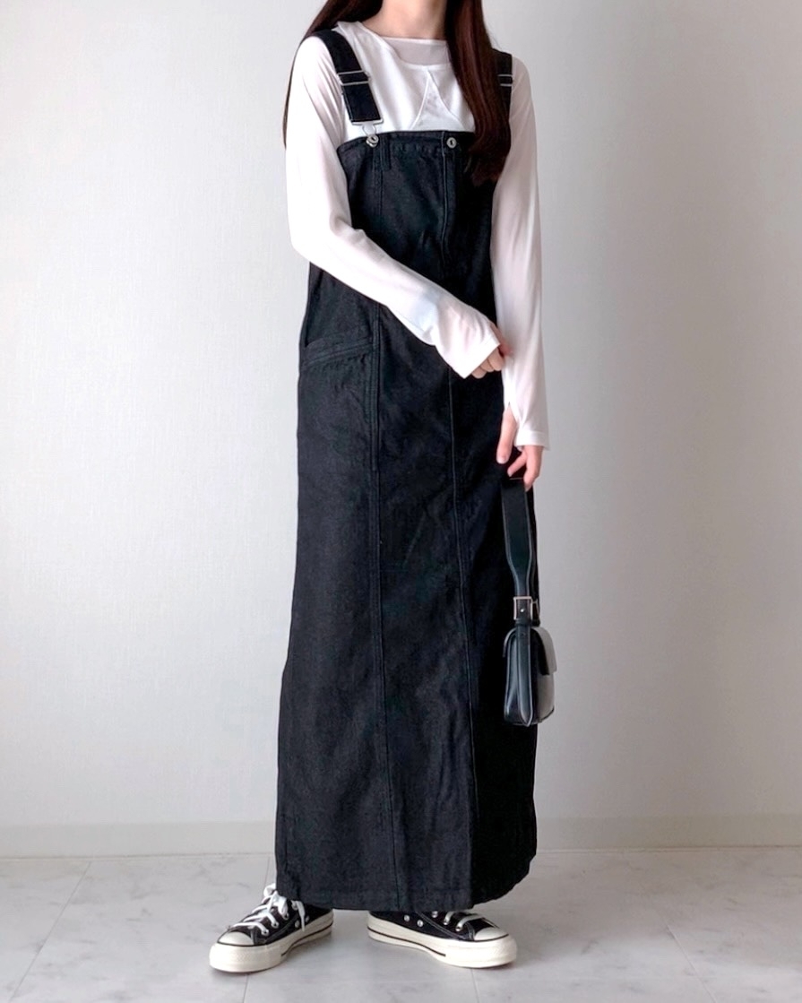 肩紐付きの黒いロングジャンパースカートを着た女性が、白い壁の前に立っている。