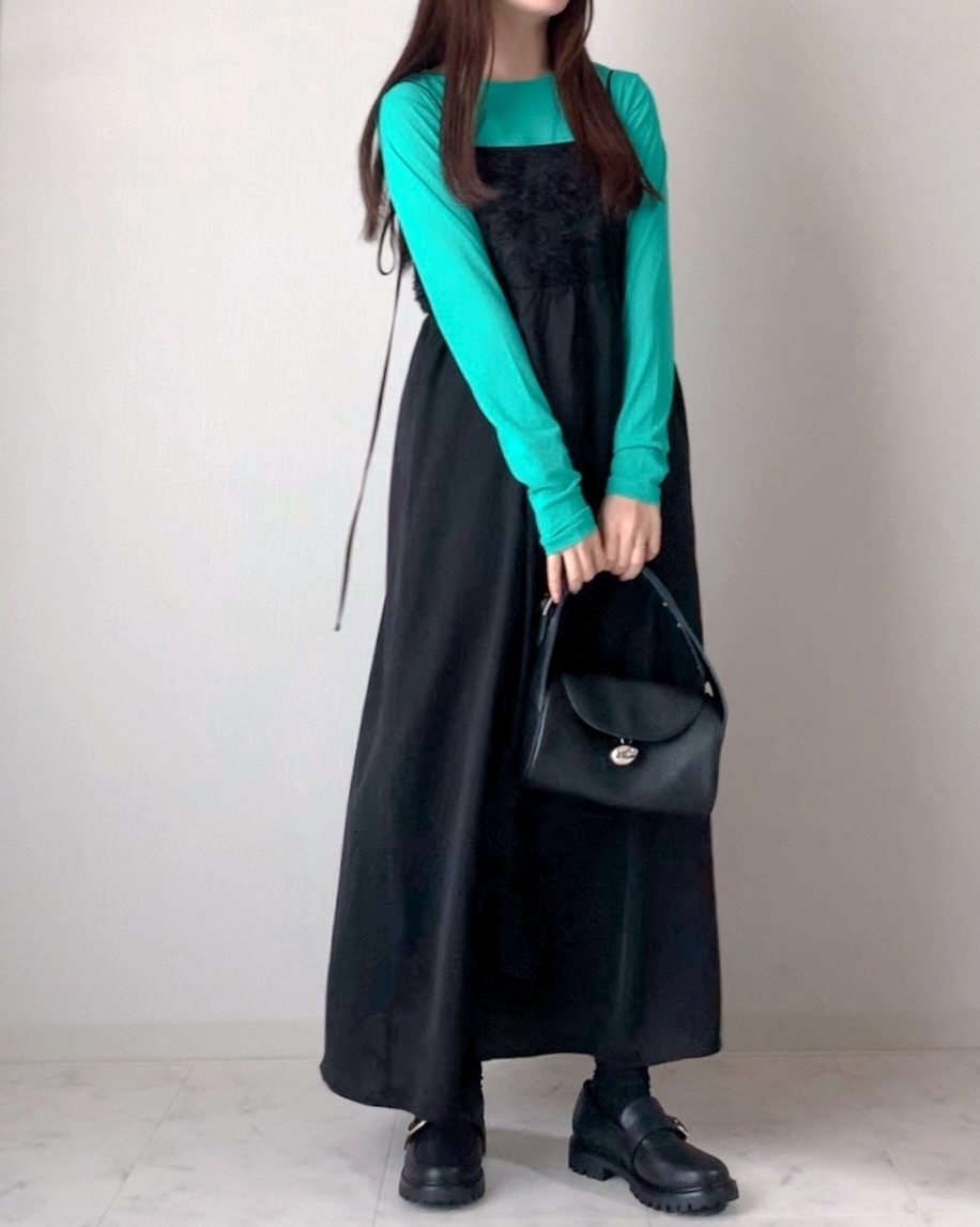 女性が長袖のトップスに黒いノースリーブワンピースと靴を合わせて立っている。黒いバッグを持っている。
