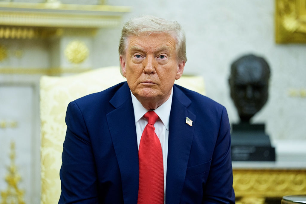A person in a suit with a red tie sits in a formal room. A bust is visible in the background