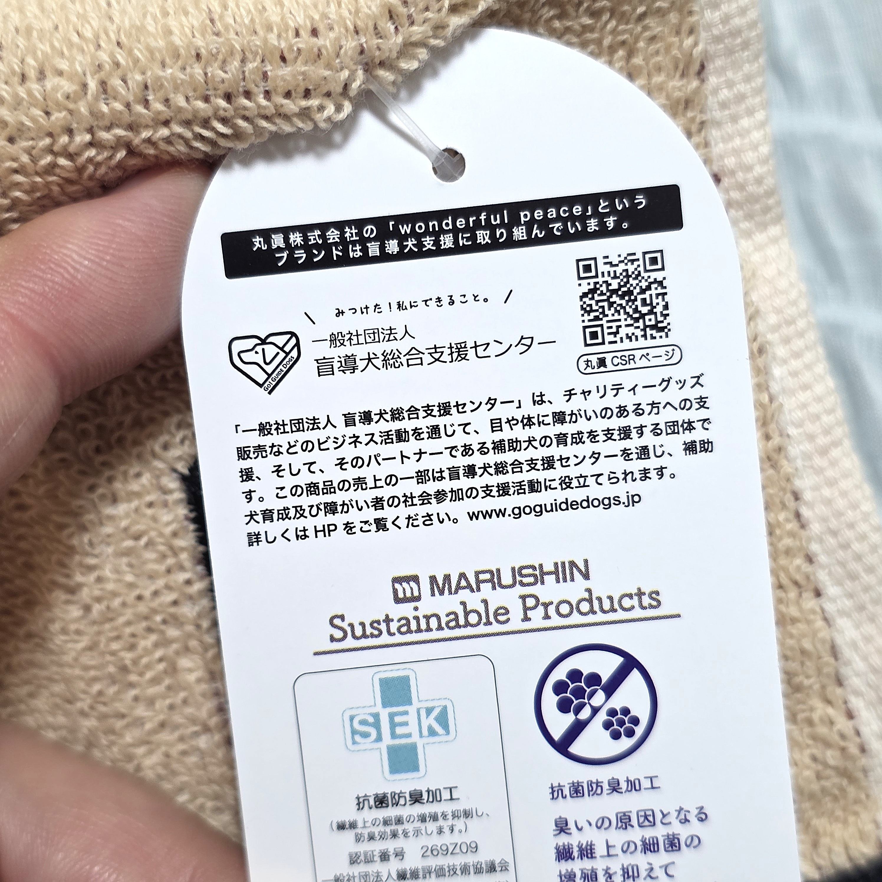 服のタグに、持続可能な製品であることと支援団体の情報が記載されている。QRコード付き。